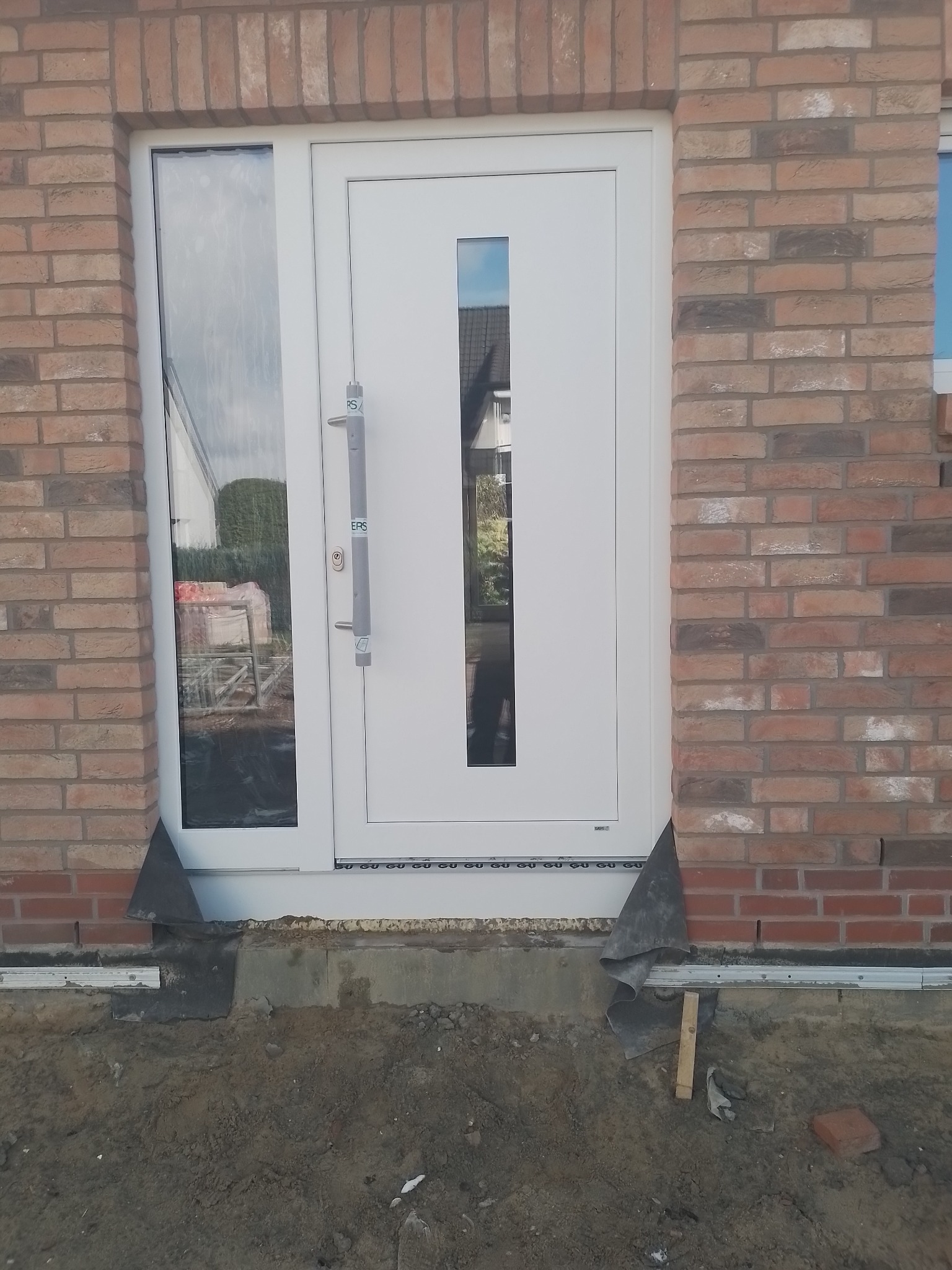 Białe drzwi wejściowe z szybą, zamontowane w murze z cegły. Widoczne elementy montażowe i fragment terenu przed wejściem. Ujęcie z bliska, skupione na detalu.