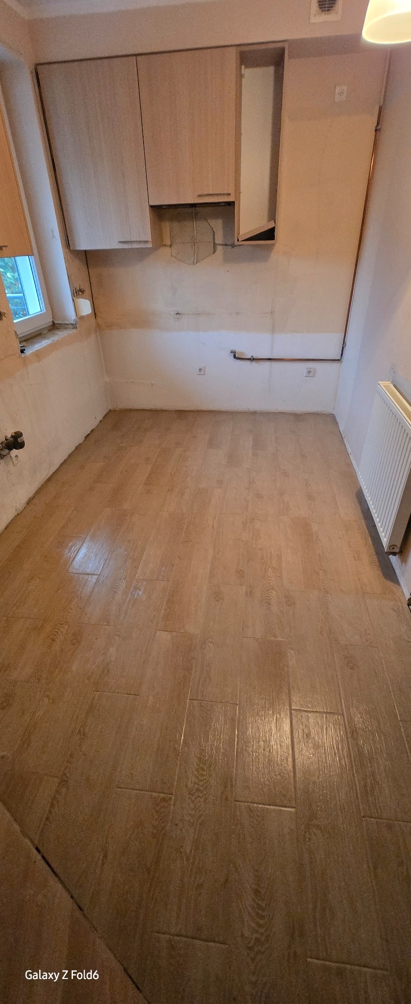 Kuchnia w trakcie remontu: położona podłoga z paneli imitujących drewno, wiszące szafki, ściany przygotowane do malowania, widoczne instalacje.