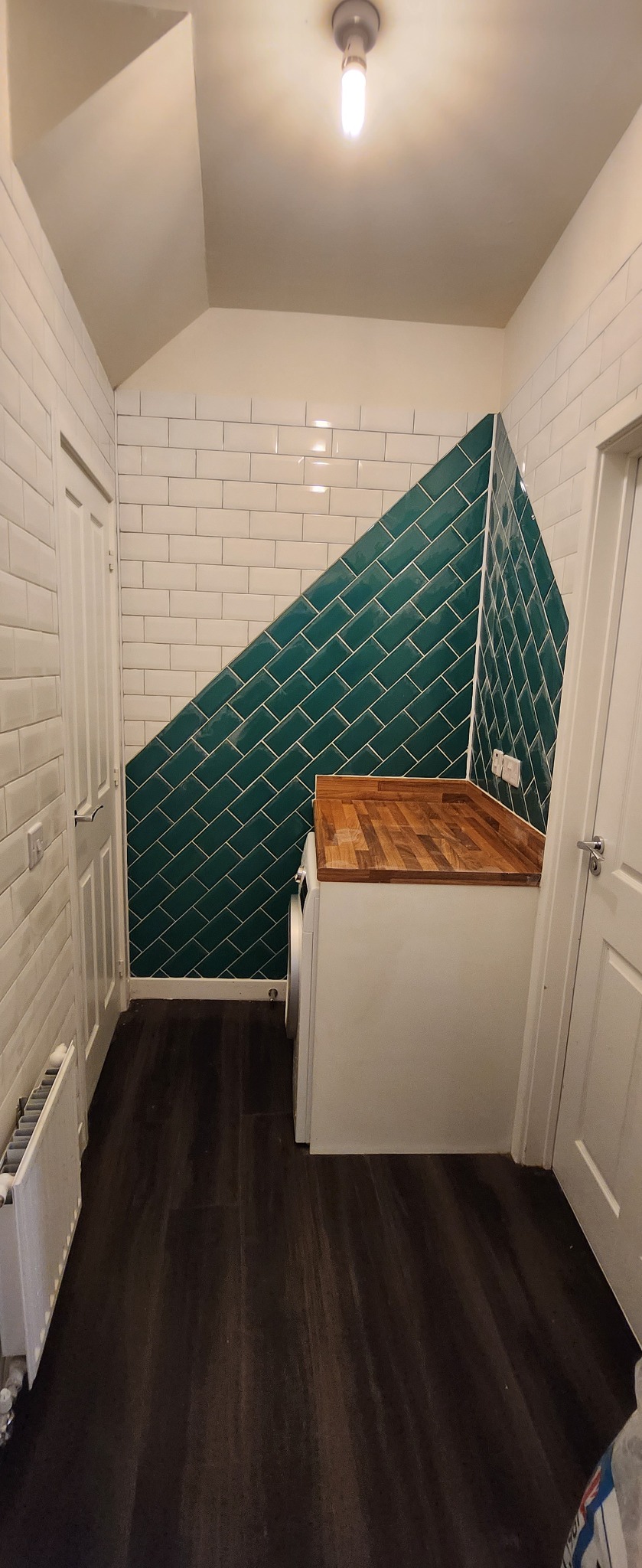 Wykończona łazienka z akcentem: białe i zielone płytki ułożone pod skosem, drewniany blat nad pralką. Ciemna podłoga i białe drzwi po bokach. Nowoczesne oświetlenie.