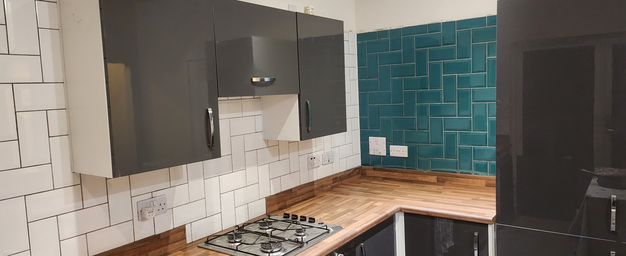 Kuchnia po remoncie: szare szafki z połyskiem, białe i turkusowe płytki ułożone w cegiełkę, drewniany blat i płyta gazowa. Widoczne gniazdka elektryczne.