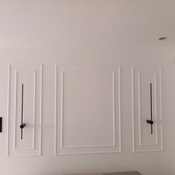 KASKADA PŁYTEK Kamil Stępień - Biała ściana z dekoracyjnymi, symetrycznymi panelami i czarnymi elementami oświetleniowymi. Widoczny fragment sufitu z otworem na lampę.