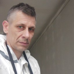 KASKADA PŁYTEK Kamil Stępień - Portret mężczyzny w białym kombinezonie ochronnym na tle szarej ściany. Widoczne zmęczenie na twarzy, sugerujące pracę fizyczną. Ujęcie z bliska.