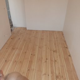 KASKADA PŁYTEK Kamil Stępień - Jasne, drewniane panele podłogowe ułożone w pokoju z białymi ścianami. Widoczny fragment ciała fotografa w dolnym rogu kadru. Naturalne oświetlenie.