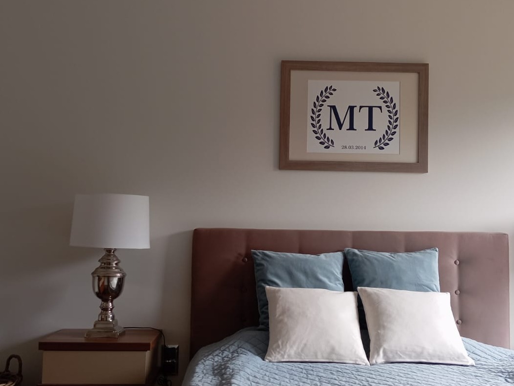 Sypialnia z łóżkiem, lampką nocną i ramką na ścianie z monogramem 'MT' i datą 28.03.2014. Neutralne kolory ścian i pościeli, miękkie światło.