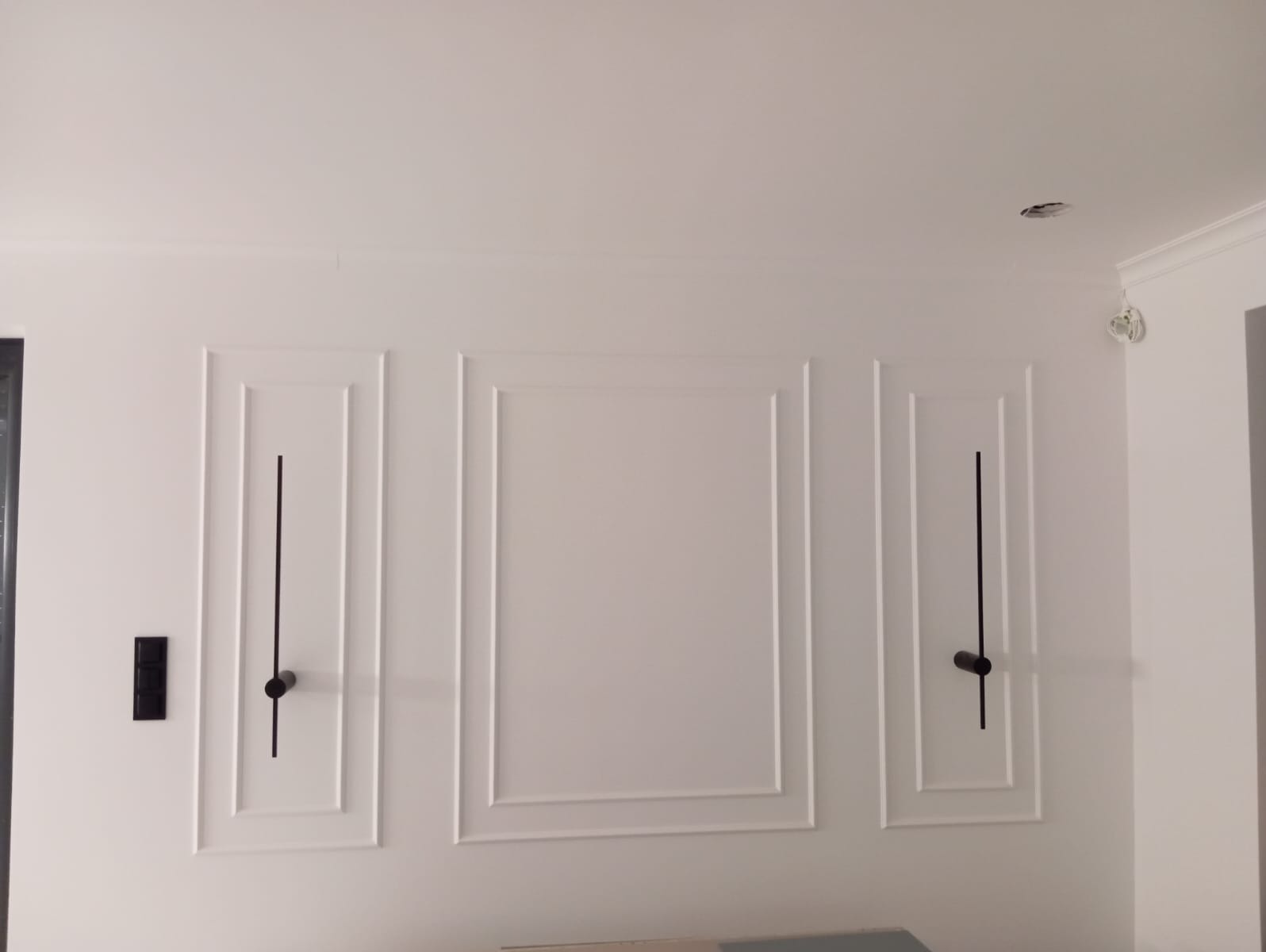 Biała ściana z dekoracyjnymi, symetrycznymi panelami i minimalistycznymi lampami. Nowoczesny design wnętrza z subtelnym oświetleniem.