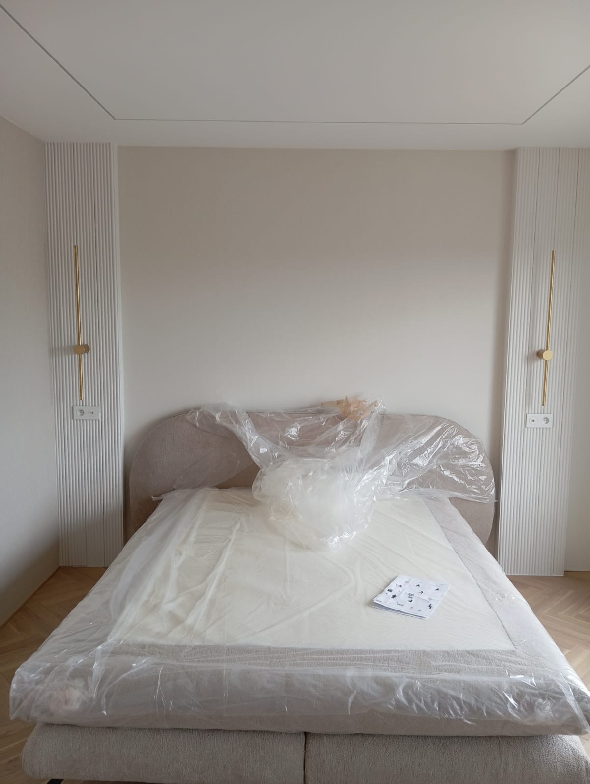Nowe łóżko z beżowym zagłówkiem, jeszcze w folii ochronnej, stoi w pokoju z panelami ściennymi i złotymi lampami. Widoczne instrukcje montażu.