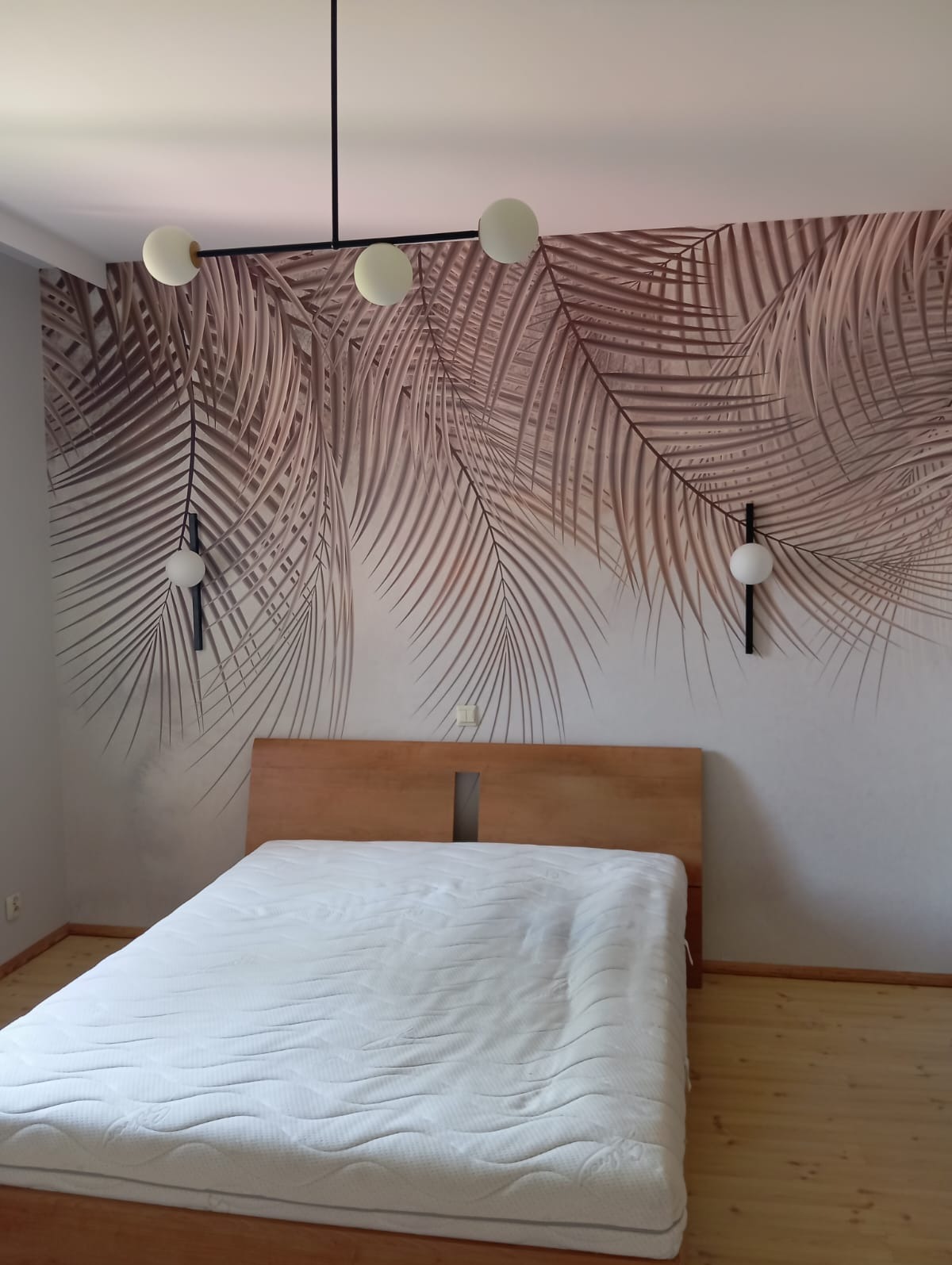 Sypialnia z łóżkiem i fototapetą w liście palmowe. Na ścianie lampy-kule, nad łóżkiem wisząca lampa. Drewniana podłoga i rama łóżka.