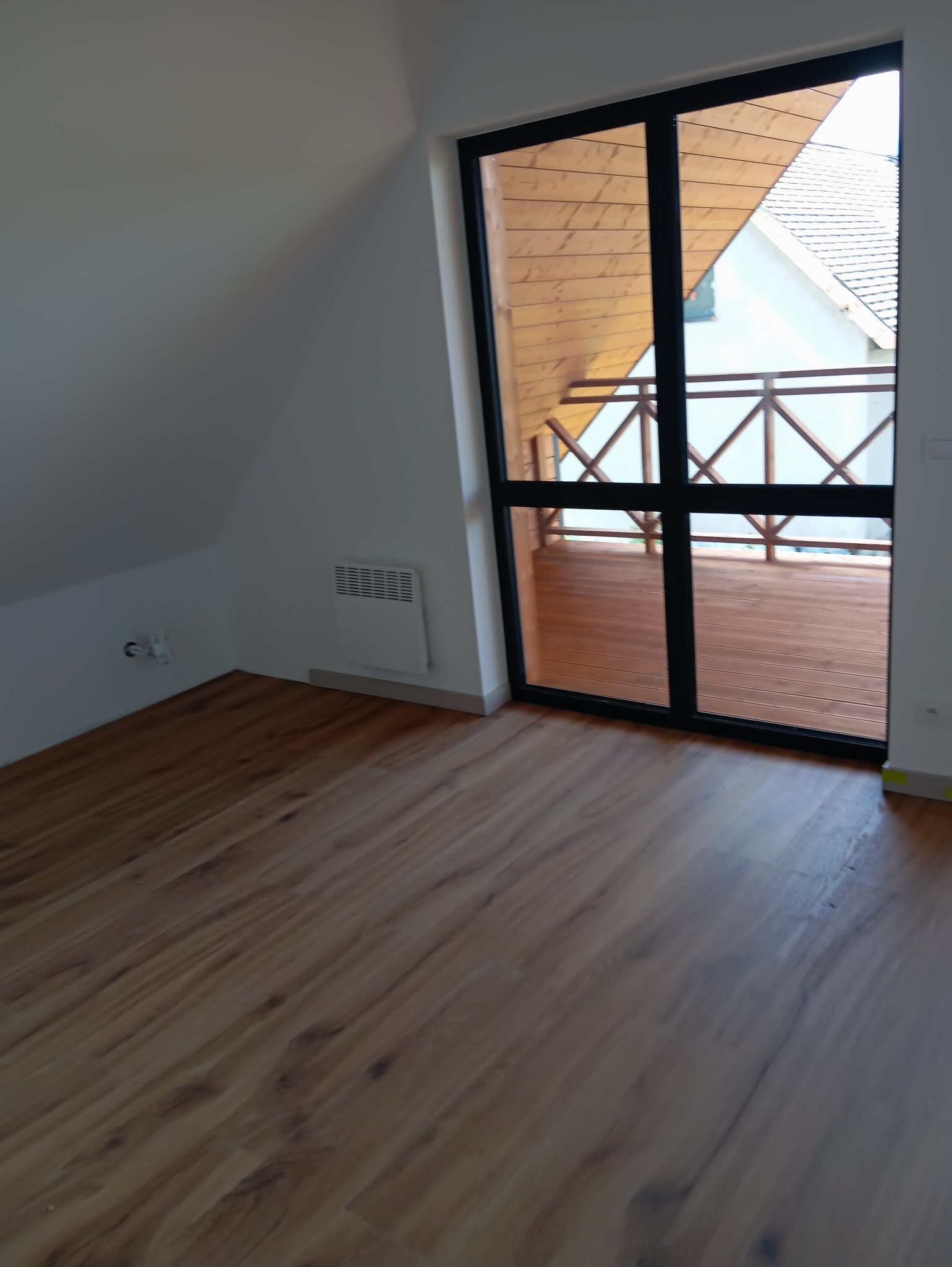 Jasne panele podłogowe w pokoju na poddaszu z oknem balkonowym. Widok na drewniany balkon i dach domu. Ściany białe, skośne. Grzejnik na ścianie.