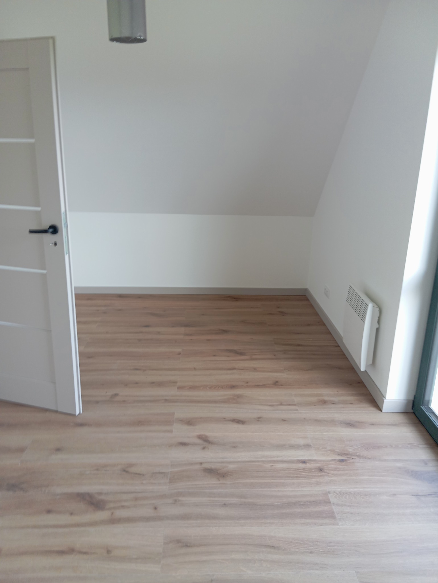 Jasne panele podłogowe w pokoju na poddaszu z białymi ścianami i otwartymi białymi drzwiami. Widoczny grzejnik elektryczny. Ukończone układanie paneli.