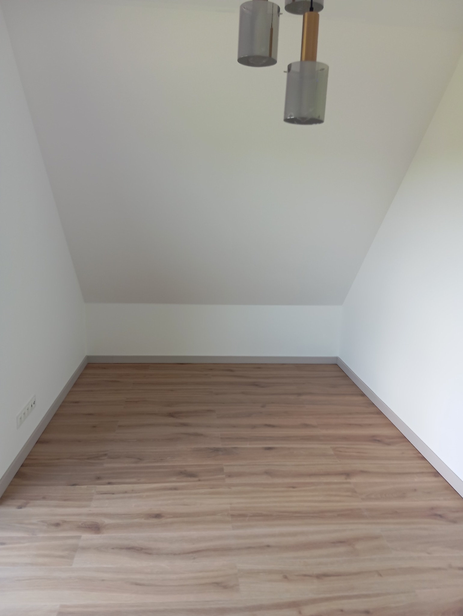 Jasne panele podłogowe w pokoju na poddaszu z białymi ścianami i nowoczesną lampą sufitową. Widok na ułożone panele, listwy przypodłogowe i gniazdka elektryczne.