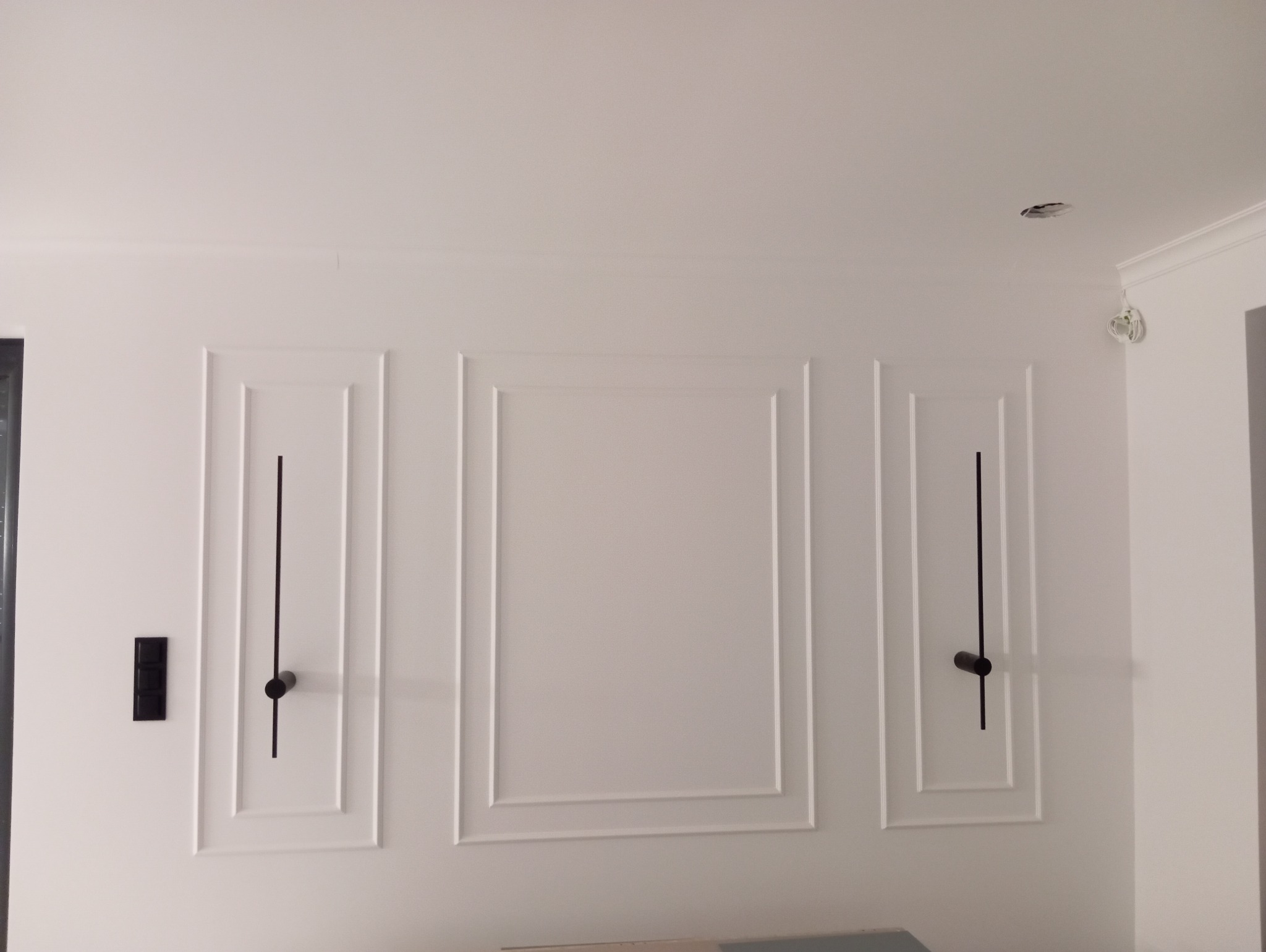 Biała ściana z dekoracyjnymi, symetrycznymi panelami i czarnymi elementami oświetleniowymi. Widoczny fragment sufitu z otworem na lampę.