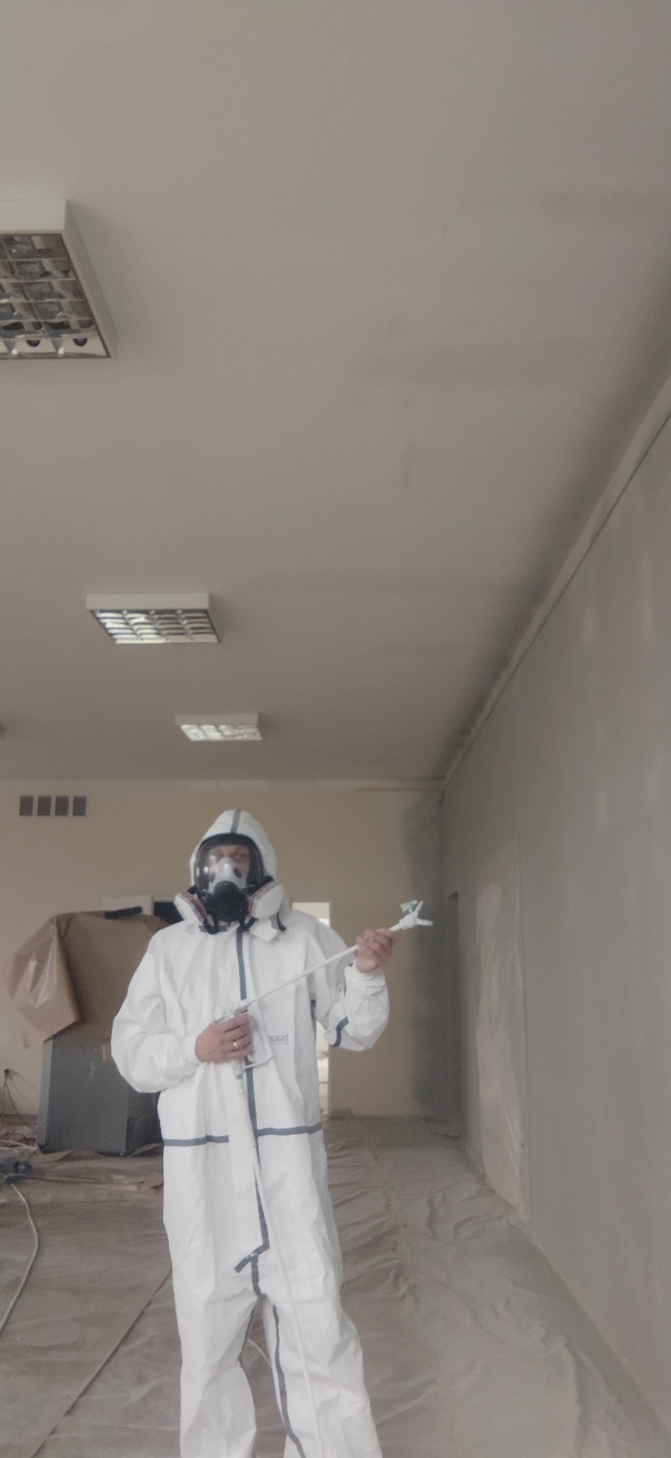 Pracownik w kombinezonie ochronnym i masce maluje ścianę natryskowo w jasnym pomieszczeniu. Podłoga zabezpieczona folią malarską. Widoczne lampy sufitowe.