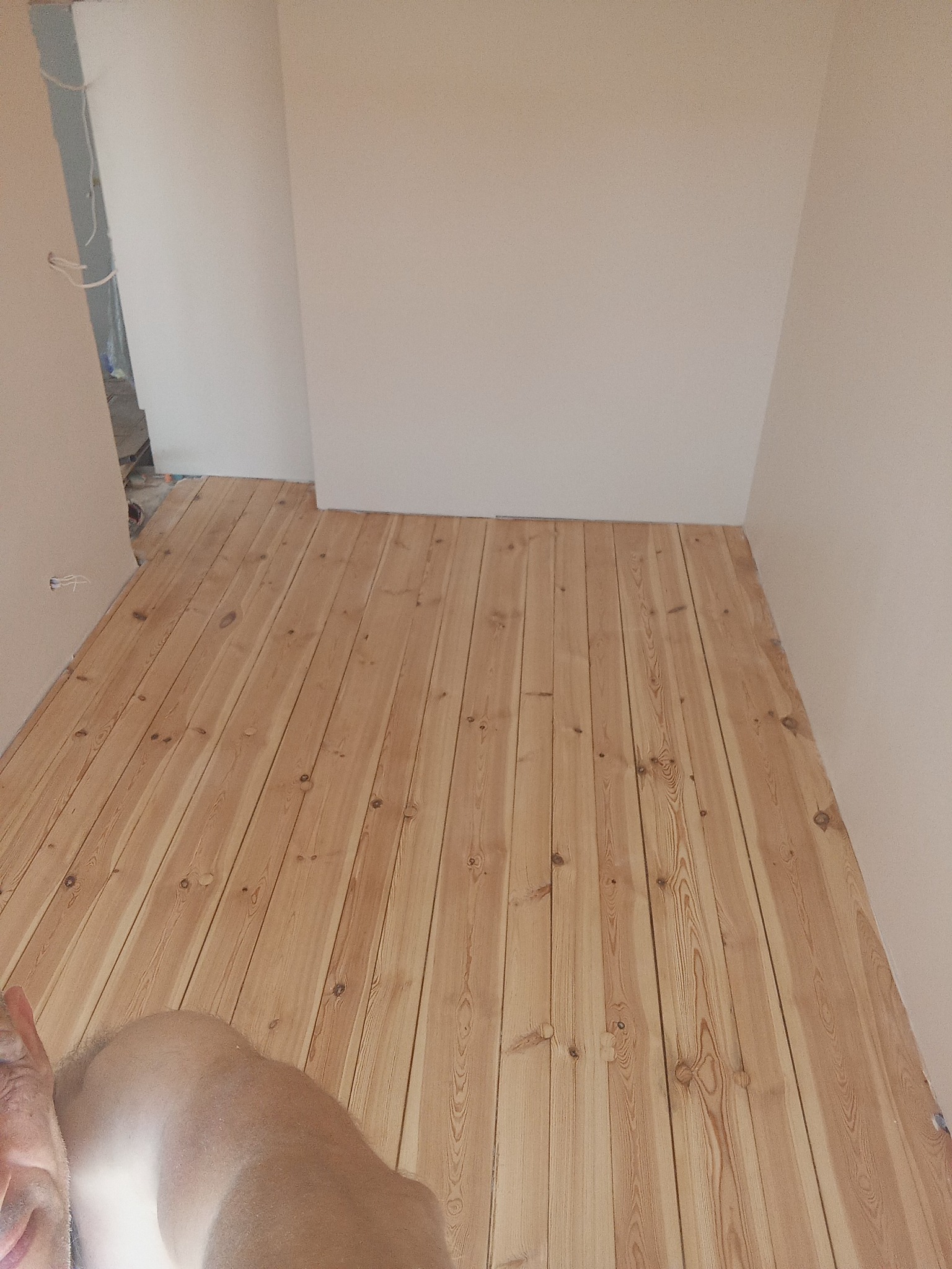 Jasne, drewniane panele podłogowe ułożone w pokoju z białymi ścianami. Widoczny fragment ciała fotografa w dolnym rogu kadru. Naturalne oświetlenie.