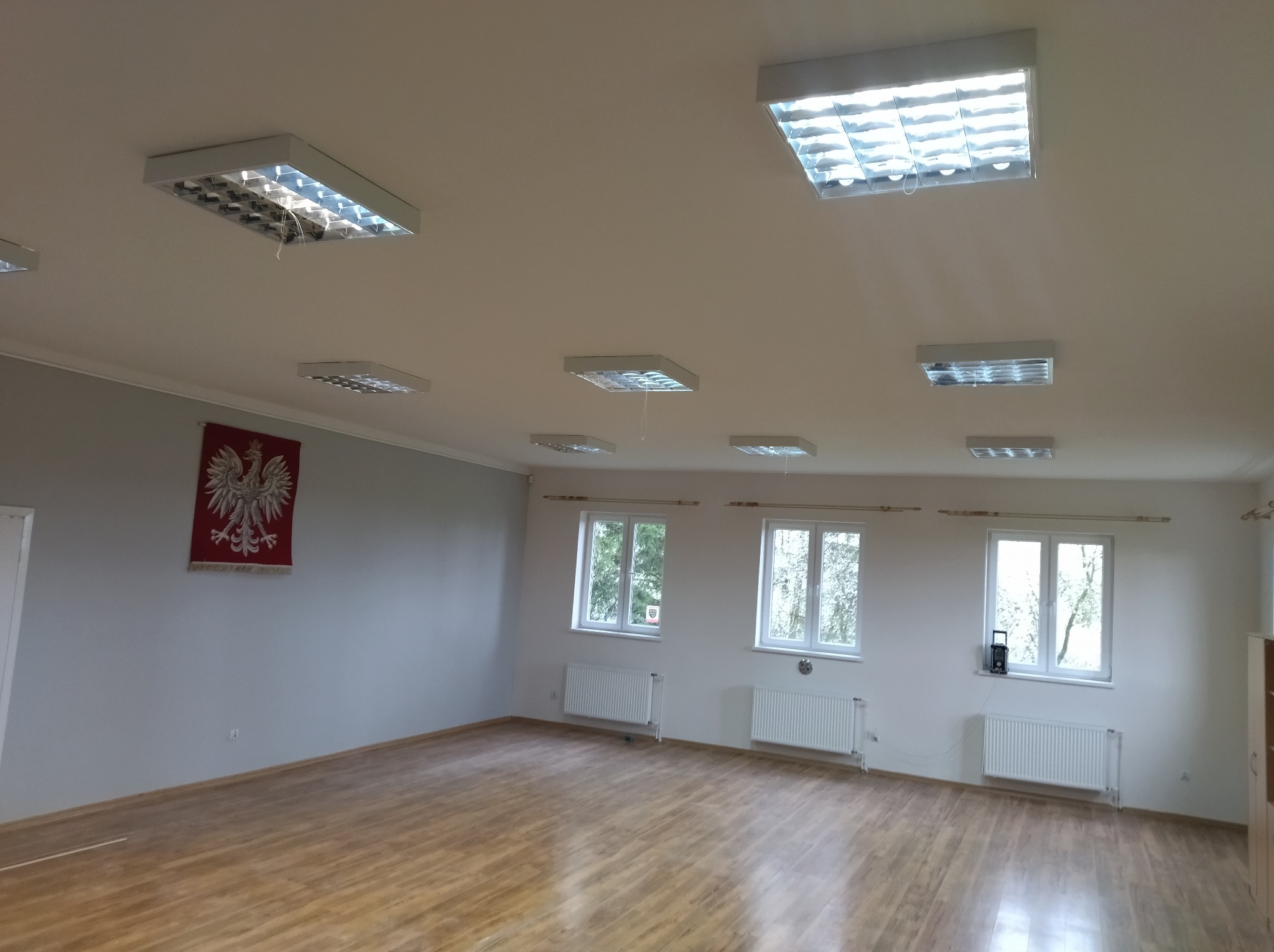 Jasne, przestronne wnętrze po remoncie: parkiet, białe ściany, okna z widokiem na zieleń, grzejniki i lampy sufitowe. Na ścianie godło Polski.