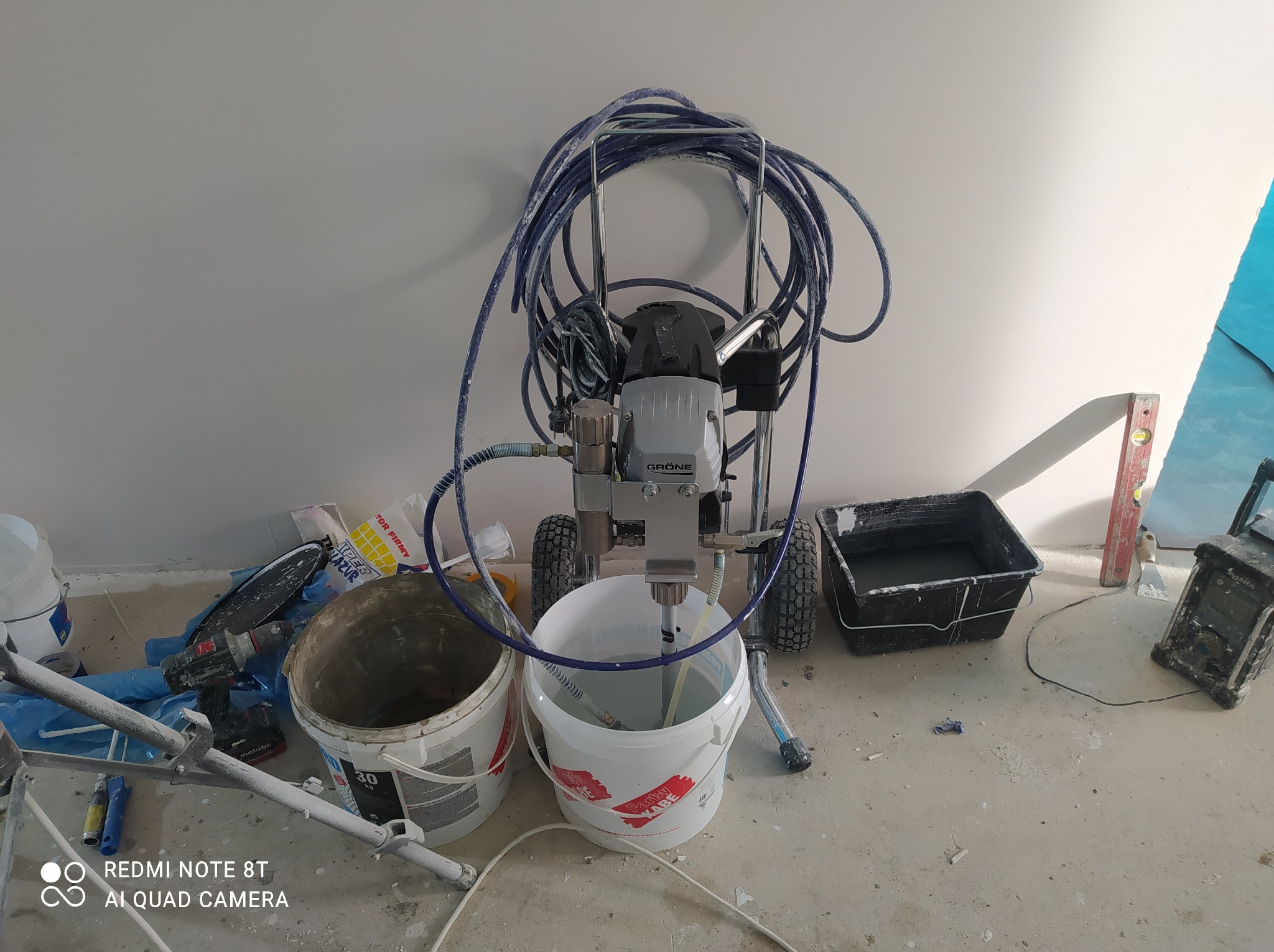 Agregat malarski Gröne z akcesoriami i wiadrami farby Kabe na podłodze w trakcie prac malarskich. Widoczny poziomica i wiertarka Metabo.