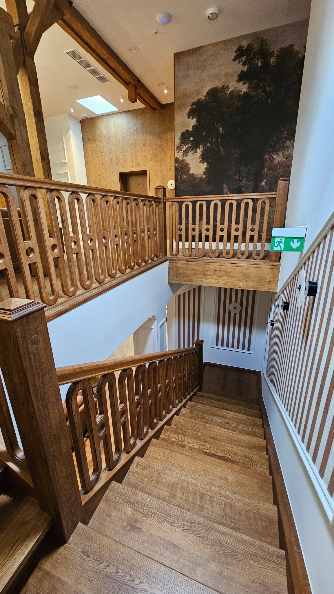 Klatka schodowa z drewnianymi schodami i balustradami o dekoracyjnym wzorze, widok z góry. Na ścianie grafika z motywem drzewa.