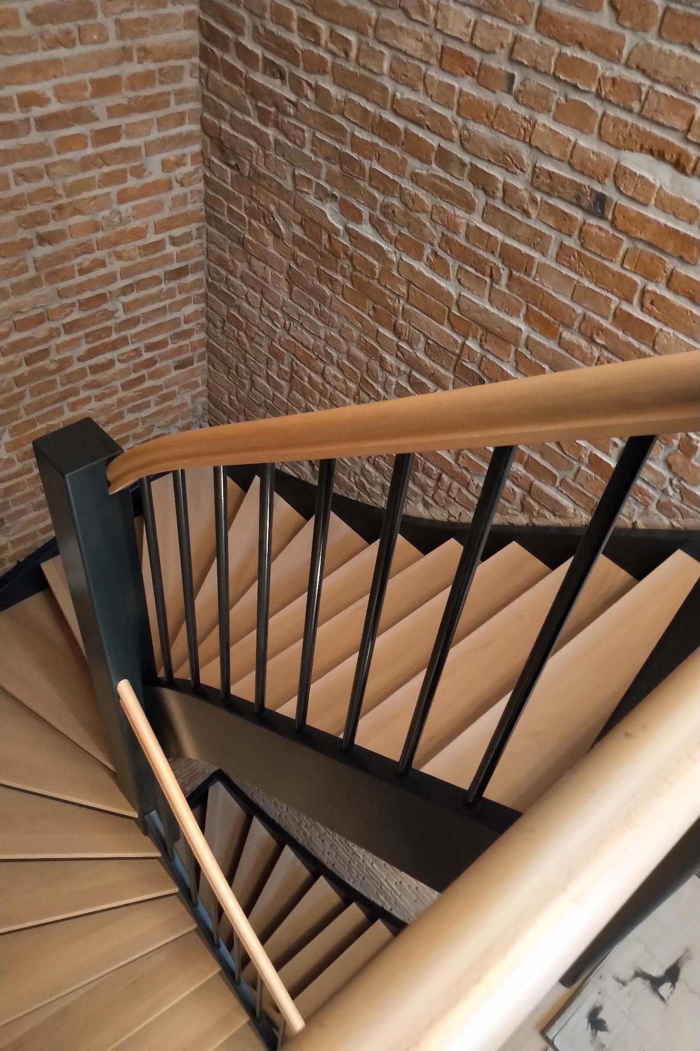 Spiralne schody z jasnego drewna z czarną metalową balustradą i drewnianym poręczą, na tle ściany z czerwonej cegły, widziane z góry.