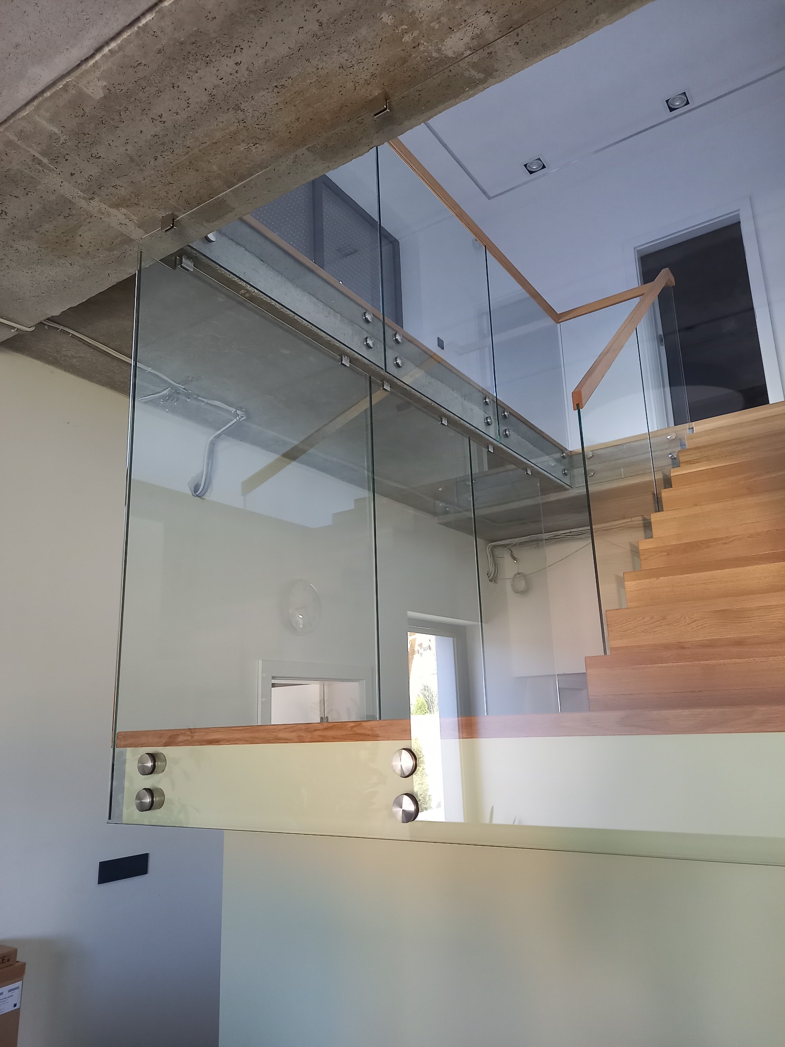 Nowoczesne schody z drewnianymi stopniami i szklaną balustradą, mocowaną na metalowych elementach. Widok z dołu, surowy betonowy sufit w tle.