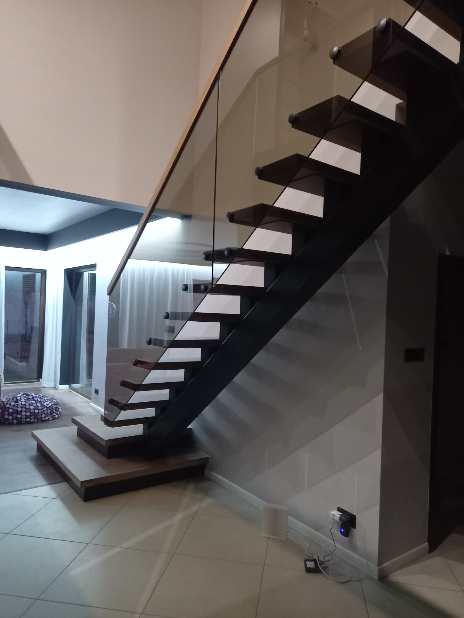 Nowoczesne schody z drewnianymi stopniami i szklaną balustradą, widoczne z dołu, z elementami oświetlenia w tle. Minimalistyczny design.