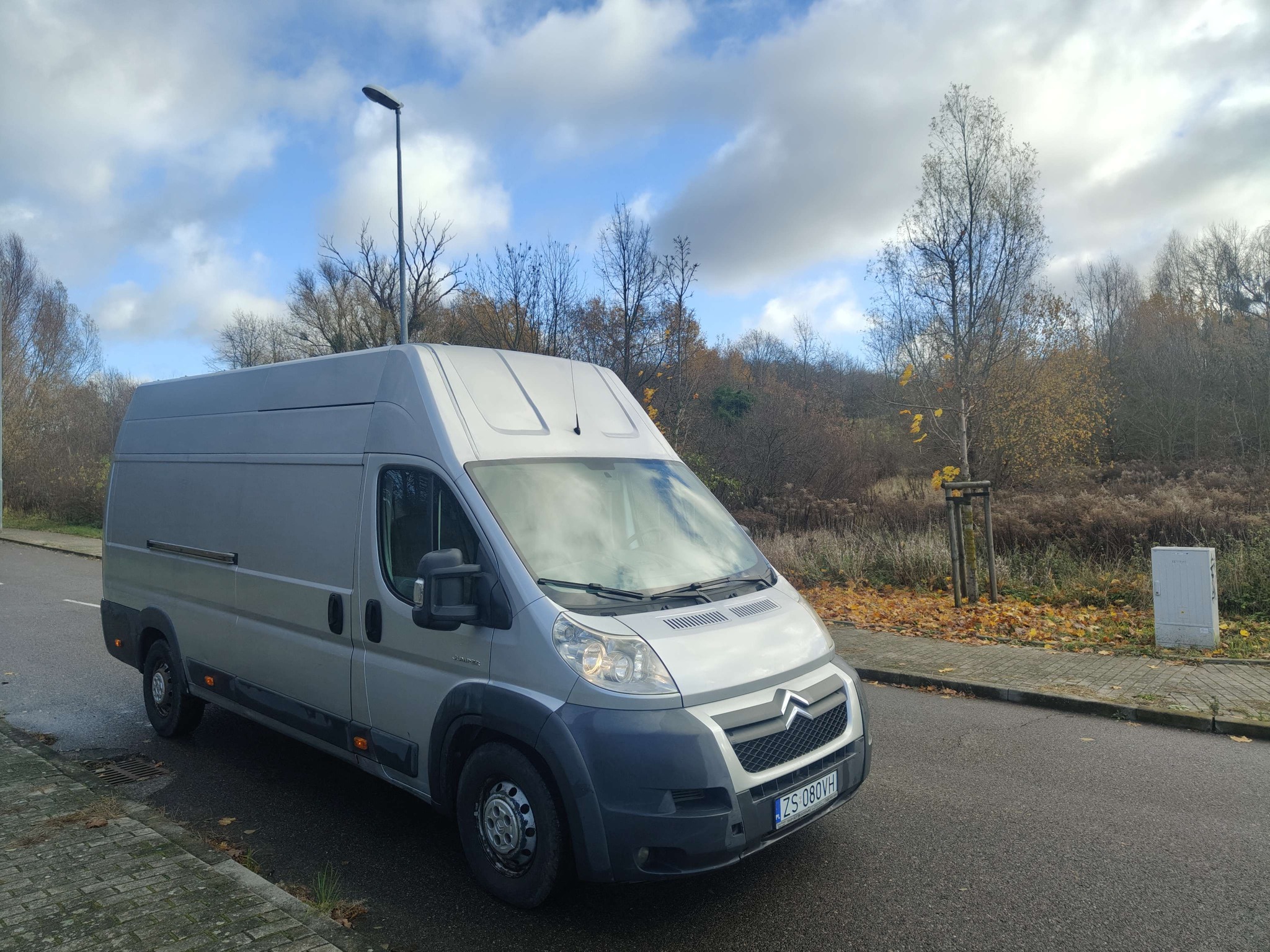 Szary bus dostawczy Citroen Jumper zaparkowany na ulicy w jesienny dzień. Widoczne drzewa w tle i błękitne niebo z chmurami. Tablica rejestracyjna ZS 080VH.