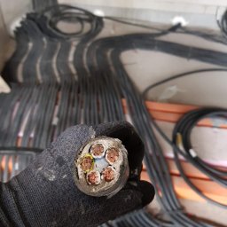 Instalacje elektryczne Damian Simiński - Przekrój grubego kabla elektrycznego z miedzianymi żyłami, trzymanego w rękawicy roboczej. W tle wiązki kabli ułożone wzdłuż ściany.