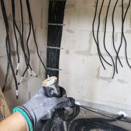 Instalacje elektryczne Damian Simiński - Rozprowadzone kable elektryczne w korytach i na ścianie z betonu komórkowego. W dłoni w rękawicy kabel z widocznym przekrojem żył.