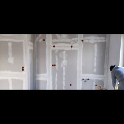 Instalacje elektryczne Damian Simiński - Ściana z płyt gipsowo-kartonowych, widoczne puszki elektryczne i instalacja wodna. W tle osoba w trakcie pracy, prawdopodobnie szpachlowanie łączeń płyt.