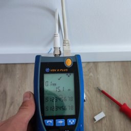 Instalacje elektryczne Damian Simiński - Pomiar długości kabla sieciowego testerem VDV II Plus, podłączonego do gniazda w ścianie. Widoczny fragment ściany, podłogi i narzędzia.