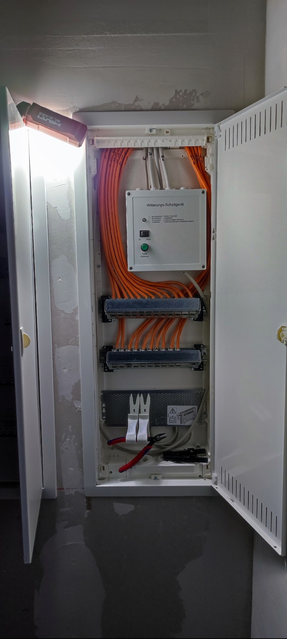 Otwarta skrzynka elektryczna z pomarańczowymi kablami, narzędzia wewnątrz, oświetlenie robocze na górze. Widoczne złącza i przewody.