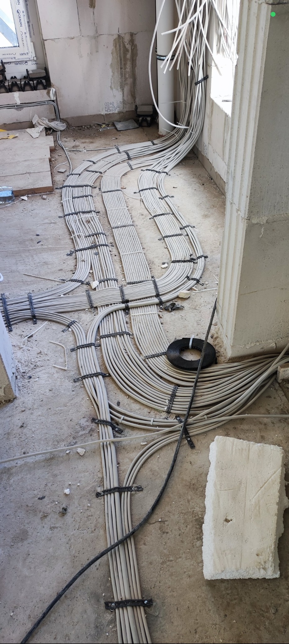 Rozprowadzone kable elektryczne w peszlach, przymocowane do podłoża w surowym stanie, widoczne elementy konstrukcyjne budynku i zwój kabla.