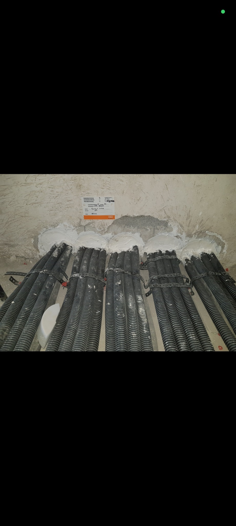 Rzędy czarnych, karbowanych rur kablowych, przymocowanych do ściany z ubytkami wypełnionymi białą masą. Widoczna etykieta z informacjami o instalacji.