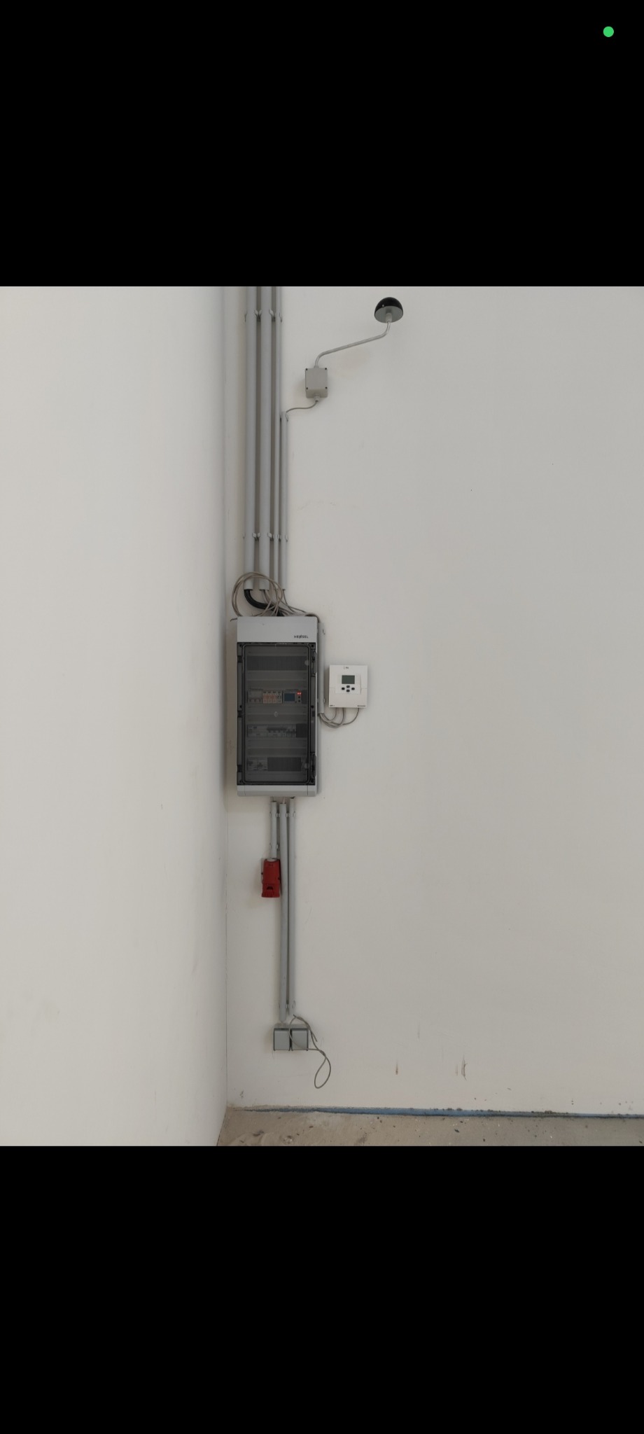 Szara skrzynka elektryczna z przewodami na białej ścianie w rogu pomieszczenia. Widoczny licznik i czerwony przycisk.