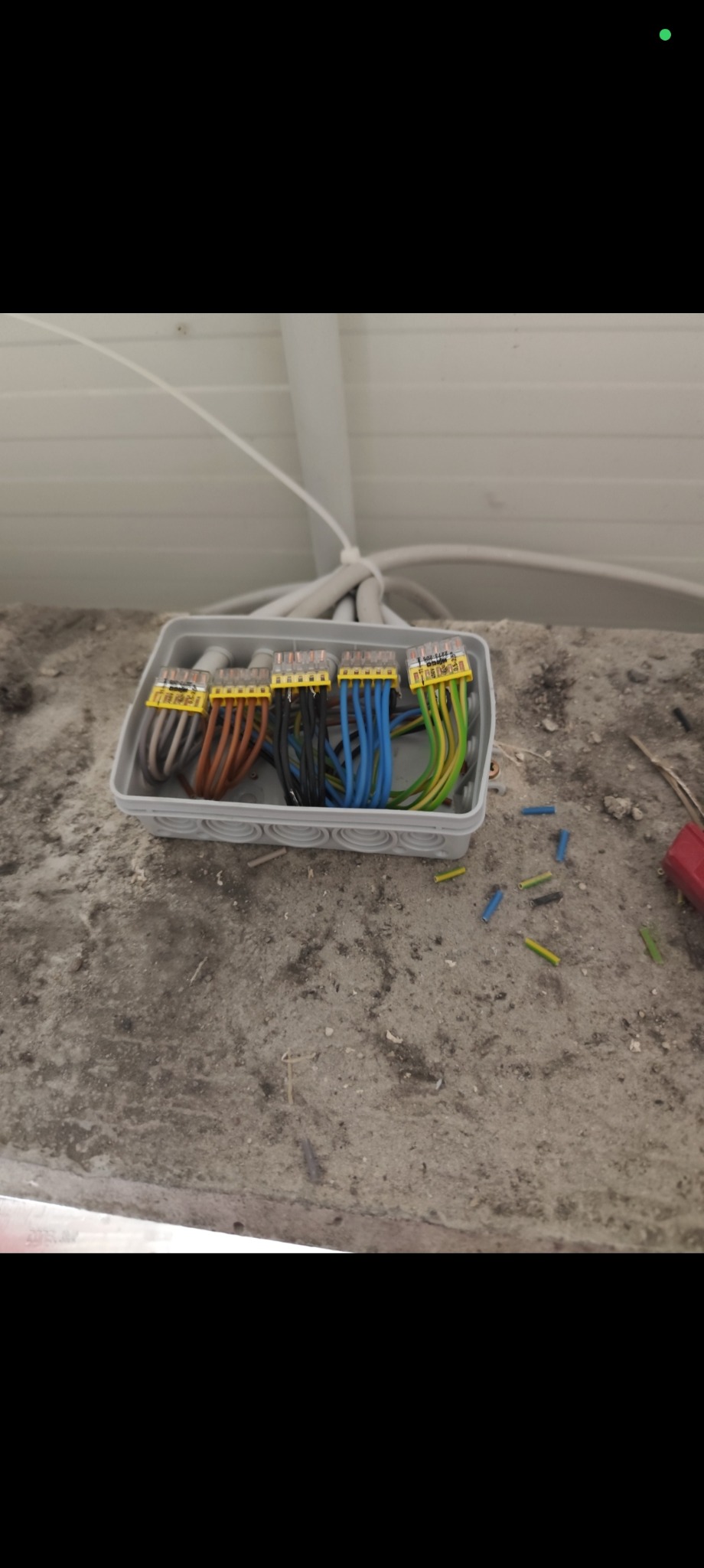 Rozdzielnica elektryczna z podłączonymi przewodami o różnych kolorach (brązowy, niebieski, czarny, zielono-żółty) na tle surowego betonu i białej instalacji.