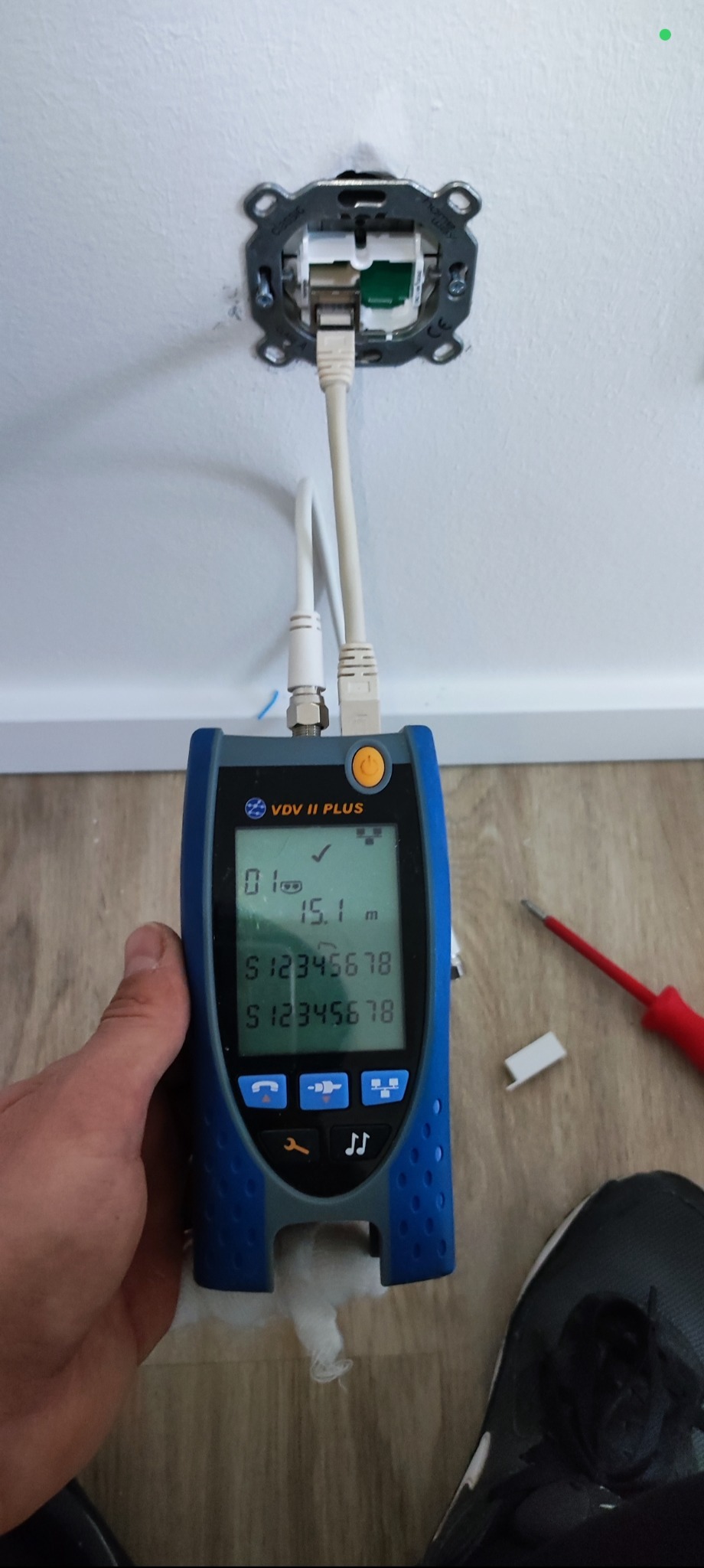 Pomiar długości kabla sieciowego testerem VDV II Plus, podłączonego do gniazda w ścianie. Widoczny fragment ściany, podłogi i narzędzia.