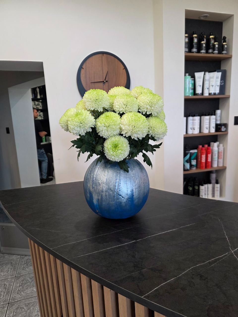 Dekoracja z białych chryzantem w niebiesko-srebrnej dyni na czarnym blacie, w tle zegar i półki z kosmetykami w salonie fryzjerskim. Sezonowa kompozycja.