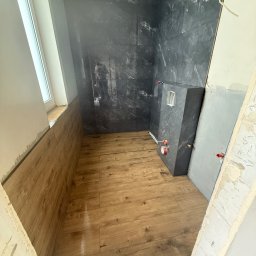 GregBud - Wnętrze łazienki w trakcie prac wykończeniowych: drewnopodobna podłoga, ściany z ciemnych płytek imitujących marmur, zabudowa podtynkowego WC. Widoczny pojemnik z narzędziami.