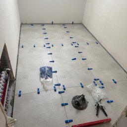 GregBud - Układanie jasnych płytek podłogowych z użyciem poziomicy i klipsów poziomujących w trakcie remontu łazienki. Widoczne narzędzia glazurnicze i instalacja ogrzewania podłogowego.