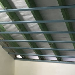 GregBud - Szkielet poddasza z drewnianymi belkami i metalowymi profilami, przygotowany pod izolację i wykończenie. Widok z dołu, surowe ściany w tle.