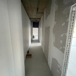 GregBud - Korytarz w trakcie prac wykończeniowych: ściany częściowo wyszpachlowane, surowy betonowy strop, widoczne profile montażowe. W tle majster przy pracy.