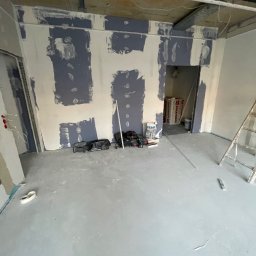 GregBud - Wnętrze w trakcie prac: ściany z częściowo nałożoną gładzią, narzędzia na podłodze, widoczne instalacje pod sufitem, drabina oparta o ścianę.