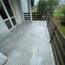 GregBud - Szare płytki na balkonie z drewnianą balustradą. Widok na zieleń. Nowoczesne wykończenie balkonu z widocznymi fugami i detalami architektonicznymi budynku.