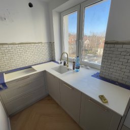 GregBud - Kuchnia w trakcie wykańczania: białe płytki ułożone do połowy ściany, szare szafki z złotymi uchwytami, biały blat, okno z widokiem na miasto. Widoczne narzędzia i materiały.