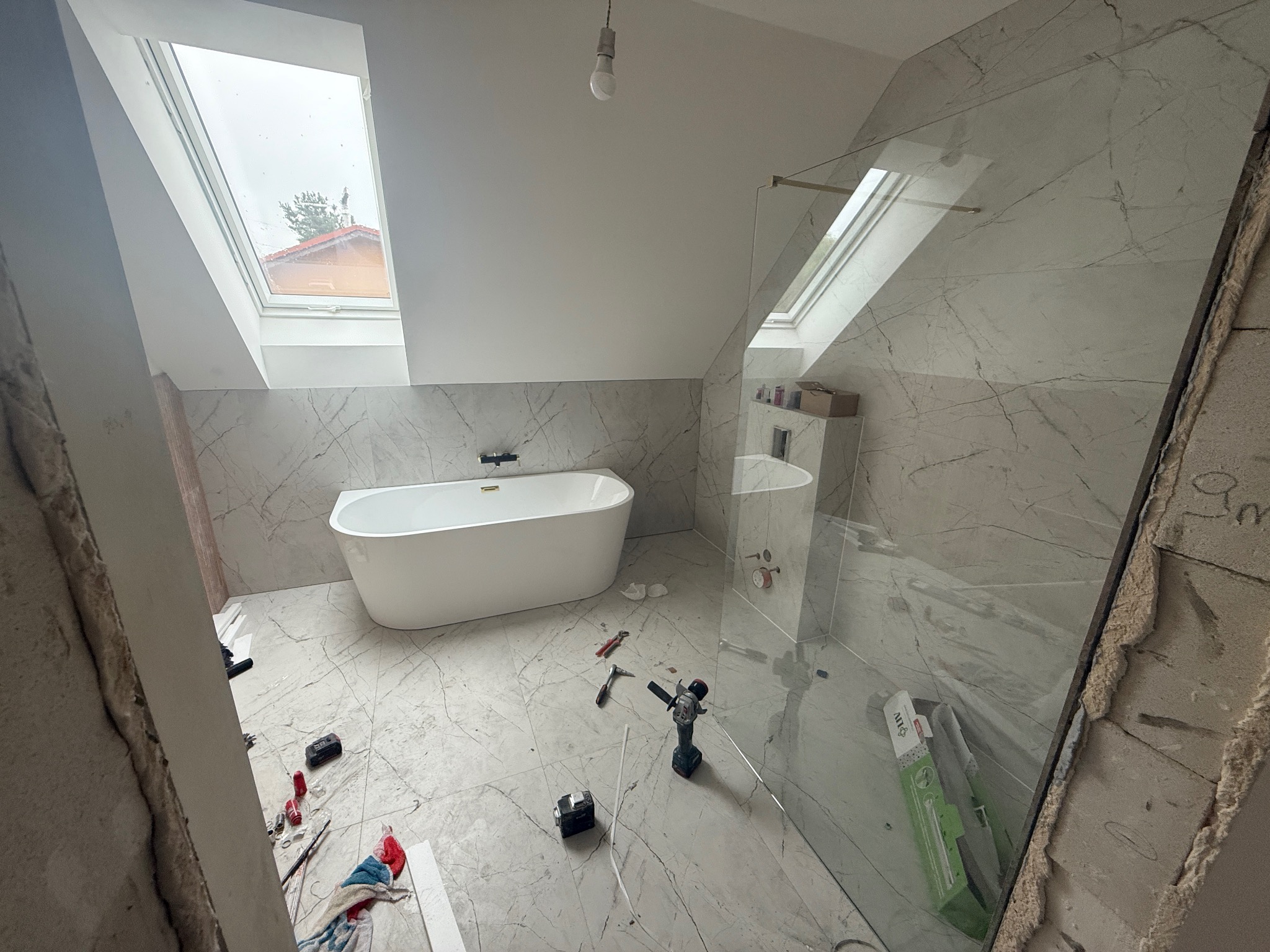 Łazienka na poddaszu w trakcie prac: biała wanna wolnostojąca, marmurowe płytki, kabina prysznicowa ze szklaną ścianką, okno dachowe. Widoczne narzędzia i materiały budowlane.