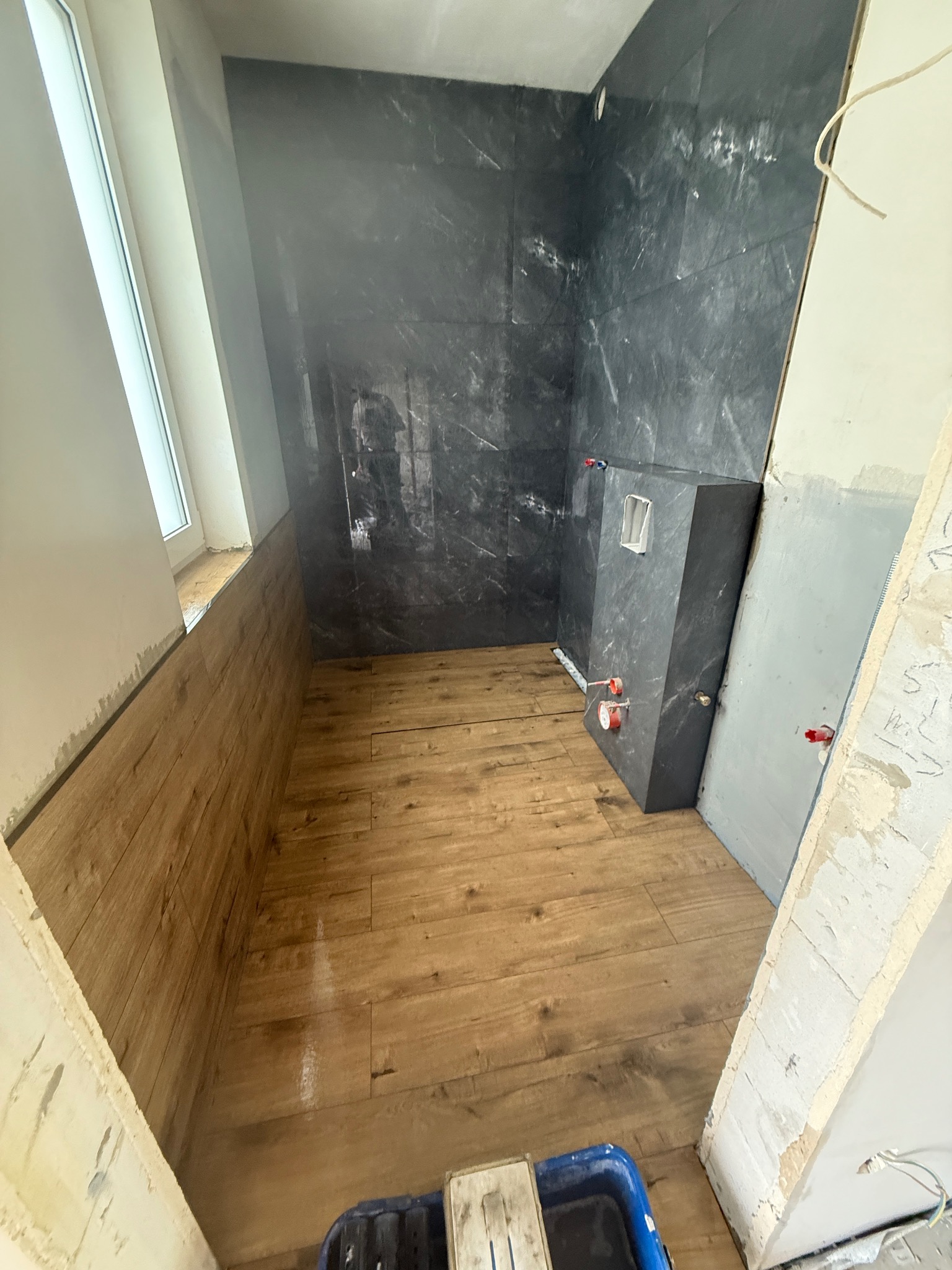 Wnętrze łazienki w trakcie prac wykończeniowych: drewnopodobna podłoga, ściany z ciemnych płytek imitujących marmur, zabudowa podtynkowego WC. Widoczny pojemnik z narzędziami.