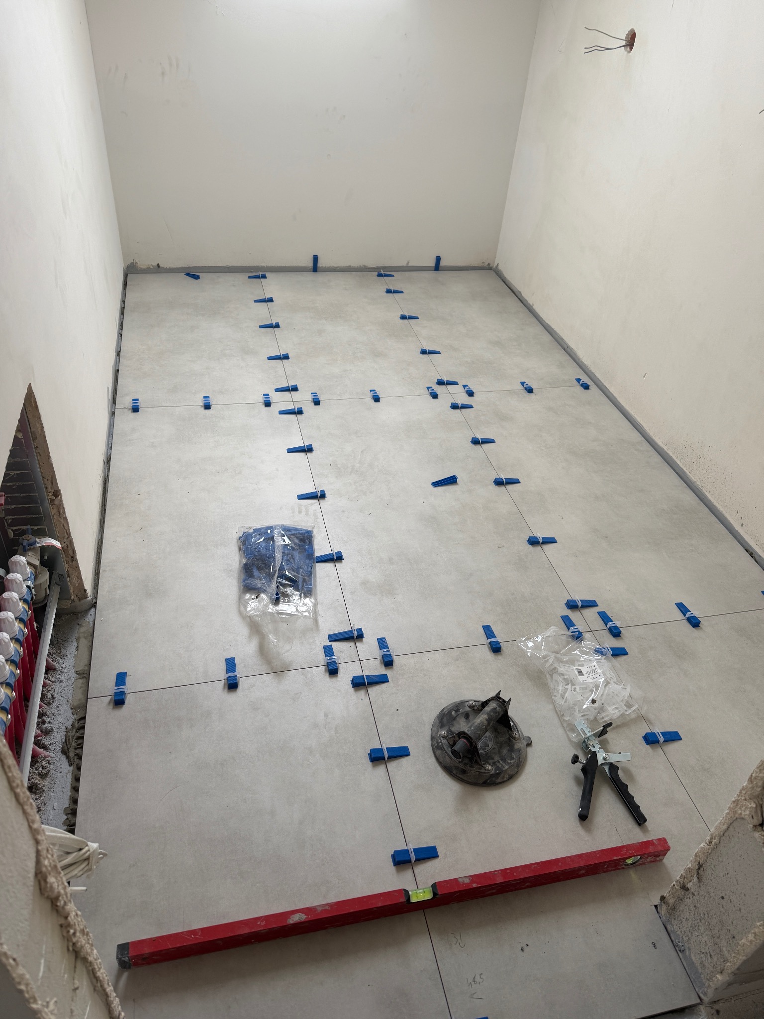 Układanie jasnych płytek podłogowych z użyciem poziomicy i klipsów poziomujących w trakcie remontu łazienki. Widoczne narzędzia glazurnicze i instalacja ogrzewania podłogowego.