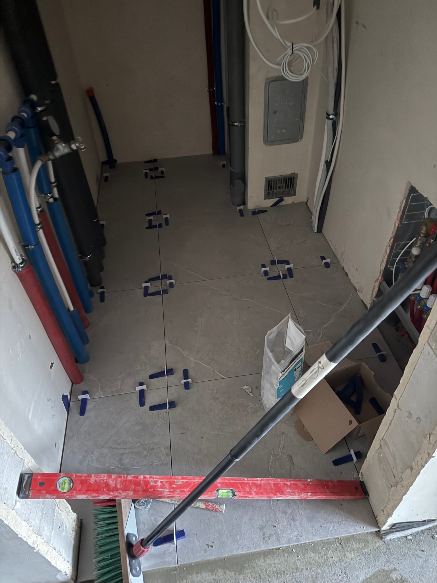 Układanie szarych płytek podłogowych w łazience, widoczne krzyżyki dystansowe, poziomica, worki z klejem i instalacja wodno-kanalizacyjna w tle.
