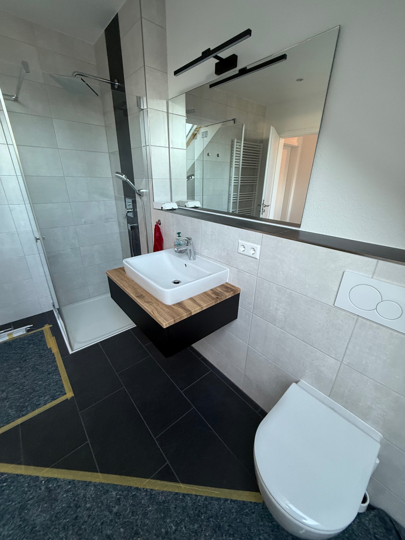 Nowoczesna łazienka po remoncie: kabina prysznicowa ze szklaną ścianką, umywalka na drewnianym blacie, czarne płytki podłogowe i biała toaleta.