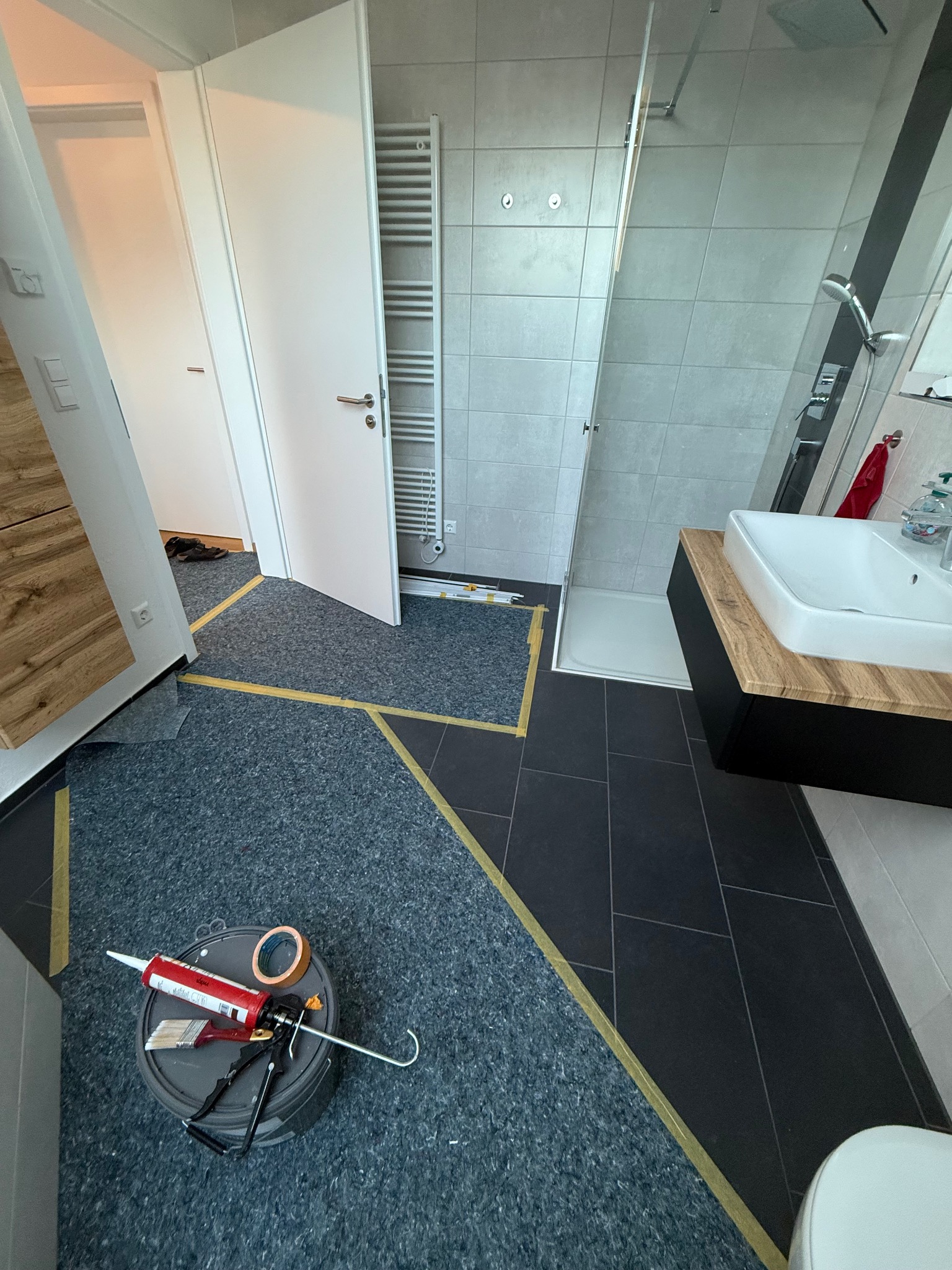 Łazienka w trakcie prac wykończeniowych: podłoga z wyznaczonym obszarem, narzędzia na wiadrze, widoczna kabina prysznicowa i umywalka z szafką.