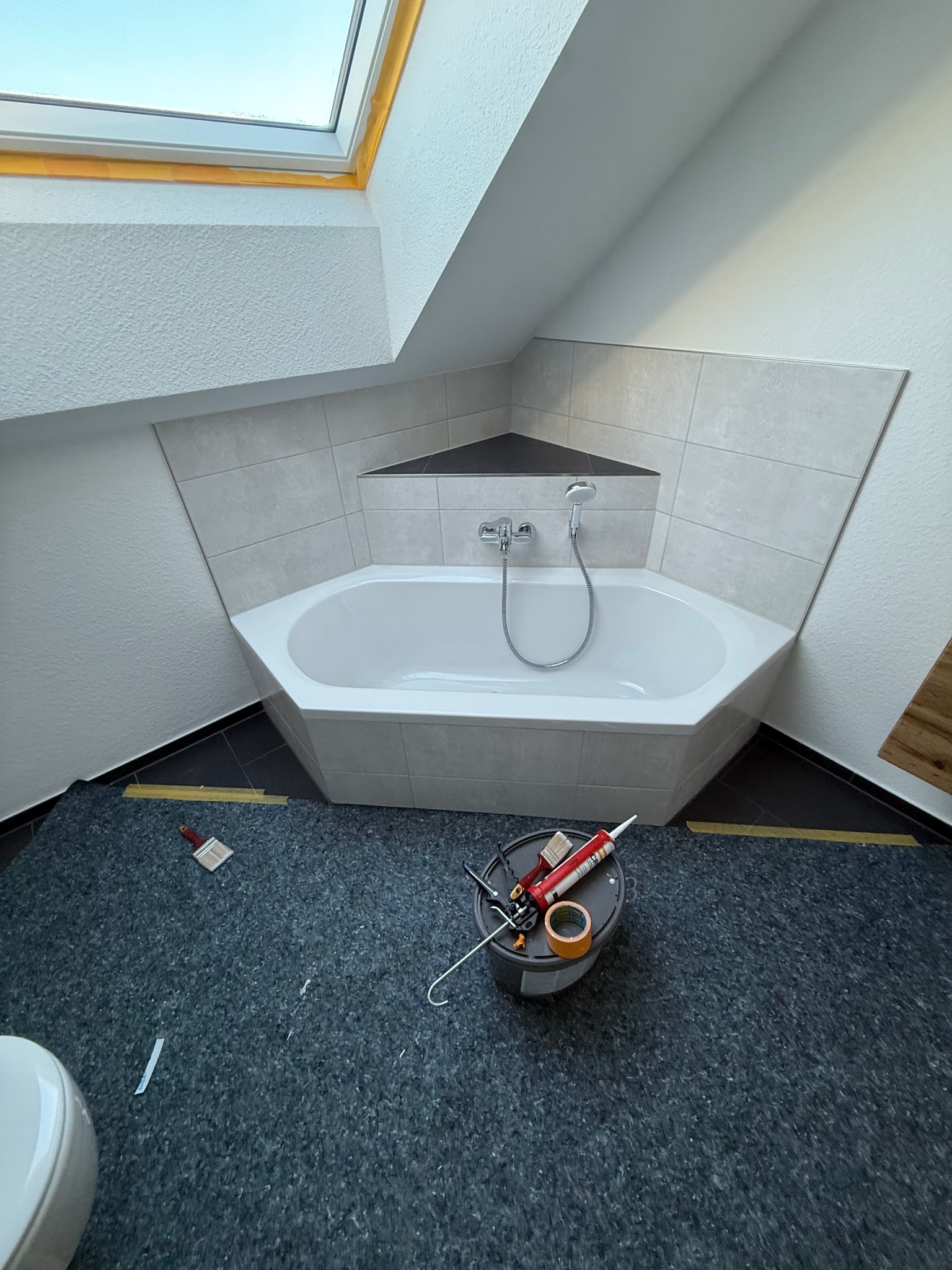 Narożna wanna w łazience na poddaszu, obudowana płytkami. Widoczne narzędzia i materiały budowlane. Okno dachowe w tle. Ukończony etap prac.