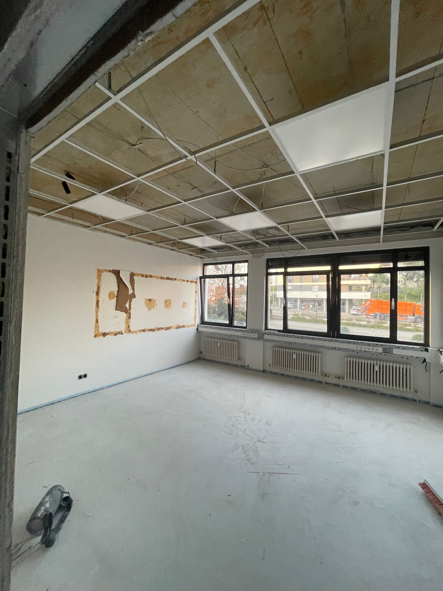 Wnętrze w trakcie prac: sufit z rastrem, ściana z fragmentami usuniętej warstwy, grzejniki pod oknami. Widoczne narzędzia i przygotowane podłoże.